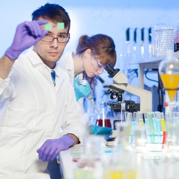 Young male researcher looking at the microscope slide in the life science (forensics, microbiology, biochemistry, genetics, oncology...)laboratory. Female asistant scientist working in the background.