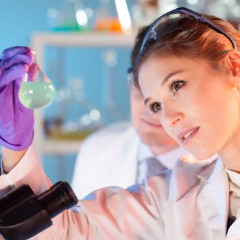 Chemical laboratory scene: attractive young student and her post doctoral supervisor scientist observing the green indikator solution color shift in glass flask.