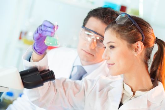 Chemical laboratory scene: attractive young student and her post doctoral supervisor scientist observing the green indikator solution color shift in glass flask.