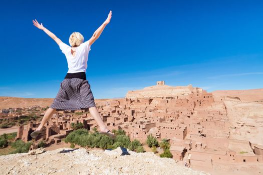 Beautiful cheerful adventurous woman jumping in front of Ait Benhaddou, fortified city, kasbah or ksar, along the former caravan route between Sahara and Marrakesh in present day Morocco.