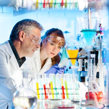 Attractive young female scientist and her senior male supervisor looking at the cell colony grown in the petri dish in the life science research laboratory (biochemistry, genetics, forensics, microbiology..)