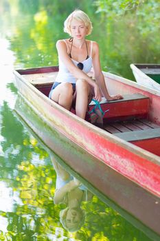 Carefree young blonde woman enjoying the sunny summer day on a vintage wooden boats on a lake in pure natural environment on the countryside.