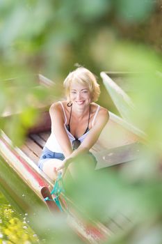 Carefree young blonde woman enjoying the sunny summer day on a vintage wooden boats on a lake in pure natural environment on the countryside.