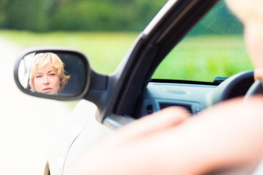 Lady driving a car in the countryside looking at the side mirror.