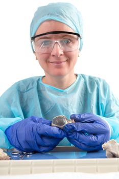 Female medical technologist or pathologist sitting in a surgical gown and cap and wearing protective goggles holding a tissue sample in her gloved hands and smiling at the camera