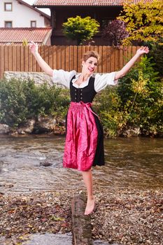 Beautiful barefoot girl in a dirndl posing at the edge of a stream with her hands raised gracefully in the air and a joyful expression