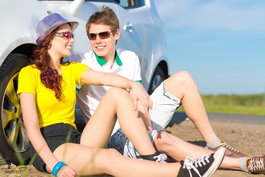 young couple sitting on the ground next to the wheel of a car, a summer road trip