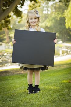 Cute Little Blonde Girl with a Bow in Her Hair Holding a Black Chalkboard Outdoors.
