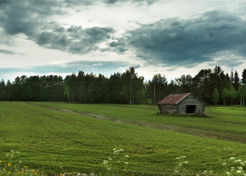Angry sky rolls in over green field with wooden barn and forest as background.