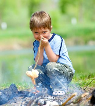 Cute boy cooking sausage by bonfire in hike.