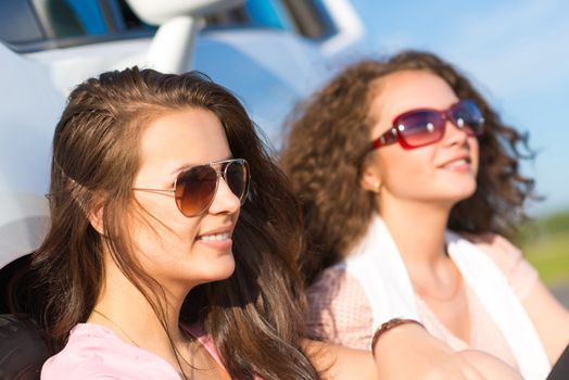 Two attractive young women wearing sunglasses, sitting next to the car