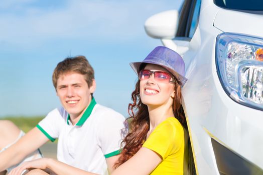 young couple sitting on the ground next to the wheel of a car, a summer road trip