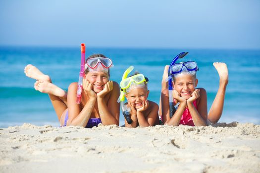 Three happy children on beach with colorful face masks and snorkels, sea in background.