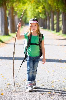 Blond explorer kid girl walking with backpack hiking in autumn trees track holding stick