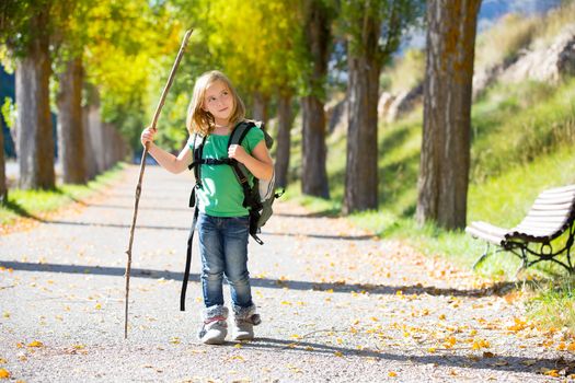 Blond explorer kid girl walking with backpack hiking in autumn trees track holding stick