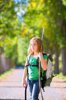 Blond explorer kid girl walking with backpack hiking in autumn trees track holding stick