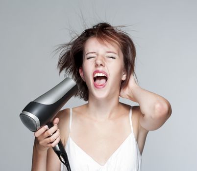 beautiful teenage girl blowing her hair with hairdryer, screaming - isolated on gray