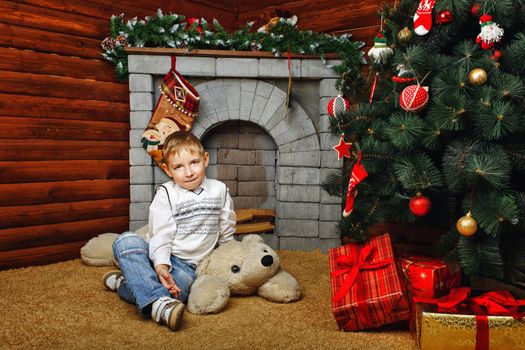 Boy sitting on teddy bear next to Christmas tree and gifts on background of fireplaces