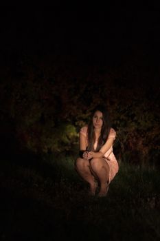 Sad young woman squatting in front of autumn bush at night