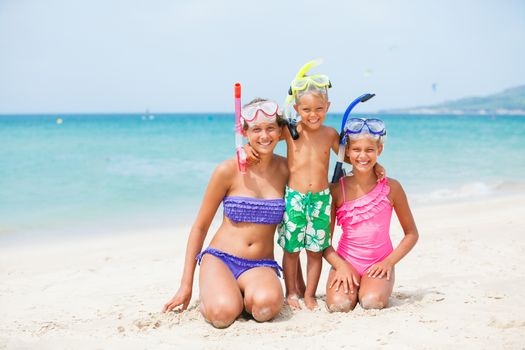 Three happy children on beach with colorful face masks and snorkels, sea in background.