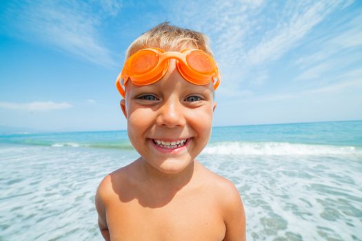Playful boy in swimming goggles on the beach with sea on background
