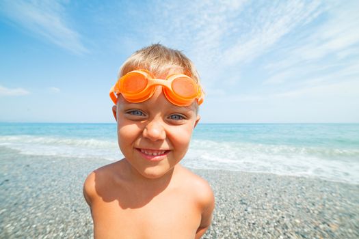 Playful boy in swimming goggles on the beach with sea on background