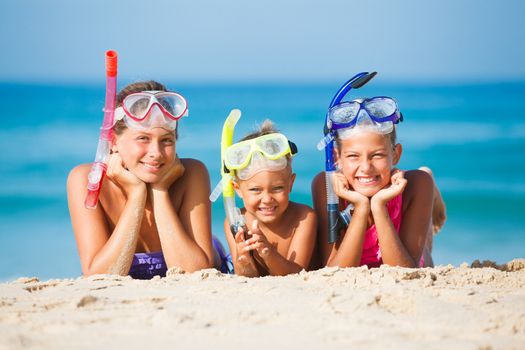 Three happy children on beach with colorful face masks and snorkels, sea in background.