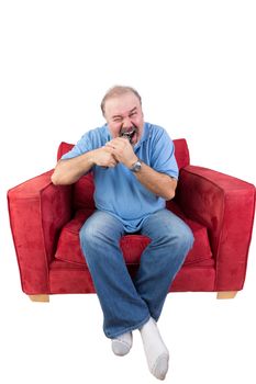 Frustrated middle-aged bearded man sitting in a red armchair biting a remote control from a television set screwing up his eyes in anger at a missed opportunity , isolated on white