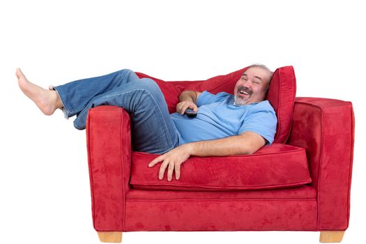 Excited man watching television laughing as he lies back barefoot on a comfortable red sofa with the remote control in his hand, isolated on white