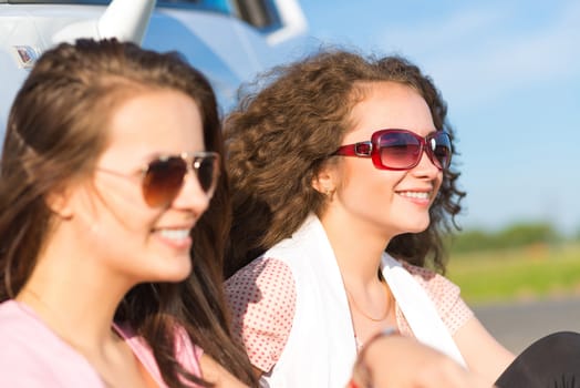 Two attractive young women wearing sunglasses, sitting next to the car