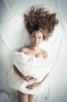 Crazy funny lady with curly hairs laying on the bed and holding pillow. Top view photo