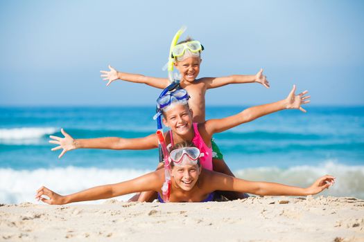 Three happy children on beach with colorful face masks and snorkels, sea in background.