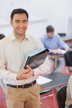 Handsome black haired teacher posing in his classroom holding some files while smiling at camera