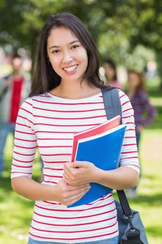 Portrait of college girl holding books with blurred students standing in the park