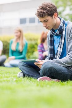 Serious young college boy using table PC with blurred students sitting in the park