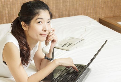 happy young asian woman working on phone and laptop in bedroom