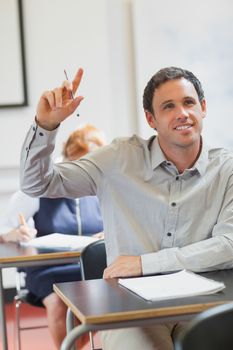 Smiling handsome mature student raising his hand while at his desk in classroom