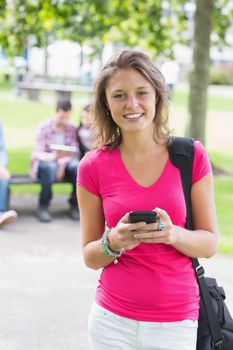 Portrait of a smiling college girl text messaging with blurred students sitting in the park