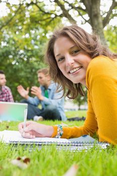 Portrait of a smiling female writing notes with two students using laptop in background at the park