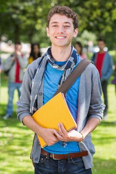 Portrait of college boy holding books with blurred students standing in the park