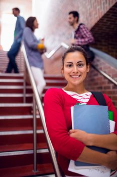 Portrait of a smiling female holding books with students behind on stairs in the college
