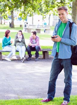 Portrait of a smiling college boy text messaging with blurred students sitting in the park