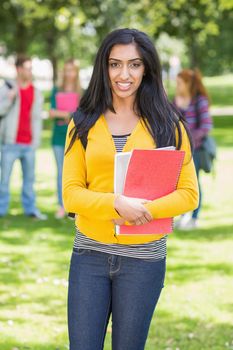 Portrait of college girl holding books with blurred students standing in the park