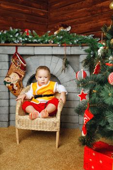 Little boy sitting in wicker chair near Christmas tree on background of fireplaces