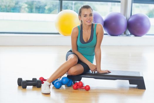 Full length portrait of a young woman sitting with dumbbells in fitness studio