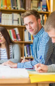 Portrait of a smiling male student with friends sitting at desk in the college library