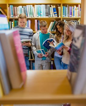 Group of four students reading book against bookshelf in the college library