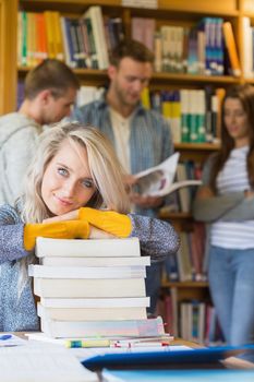 Portrait of a smiling female student with stack of books while others in background at the college library