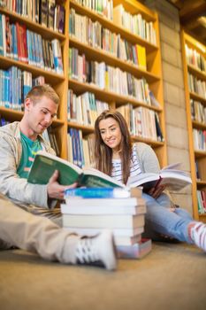 Two young students reading books against bookshelf while sitting on the library floor