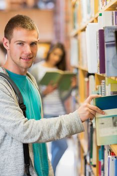 Portrait of a young smiling student selecting a book from bookshelf in the library